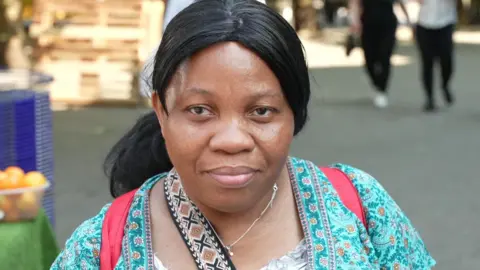 Ann Marie has black hair, which is tied back, and is wearing a blue patterned top and a red rucksack. She is standing beside a market stall.