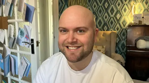 BBC / Grace McGrory Harry Finnigan, smiling at the camera. He is wearing a white top and is bald and has a beard. He is sitting in a living room. There is an armchair behind him, and a brown wood piano. To the left hand side is a door, filled with cards that have been stuck on it celebrating a new baby boy. 