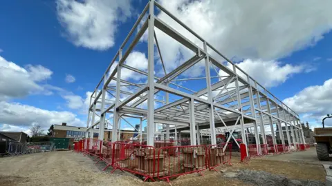 A large white metal steel structure supported by multiple columns in a building site. Out of view but beneath the structure is a large concrete swimming pool shell.
