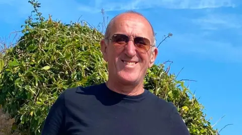 Ken Fording The image shows a smiling man wearing sunglasses, a black T-shirt and denim shorts stands outdoors beside a stone wall, with green foliage and a bright blue sky in the background.