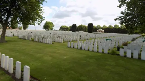 A general view picture of the New Irish Farm cemetery near Ypres. Many rows of white gravestones and lawns are surrounded by trees. 