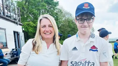JACKIE STORER A woman and a boy dressed in cricket whites stand beside a pitch and clubhouse. The woman has long blonde hair and the boy is wearing a blue cap and glasses.