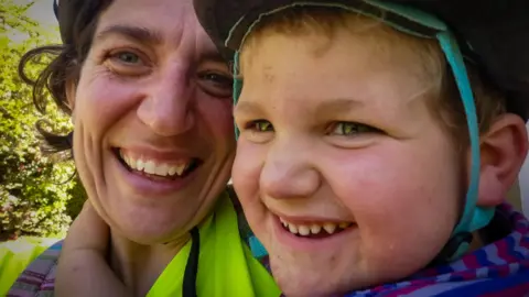 Family photo Ivor and his mum embrace for a selfie. both are smiling broadly and Ivor has his hand around mum's neck. They are wearing bike helmets and there is greenery in the background.