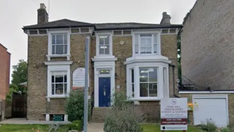 A Google screen grab of a brown brick building with large windows and a blue front door. The signage for the school is in a front garden on the grass.