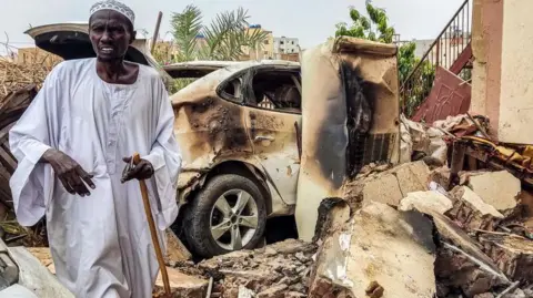 A man inspects damage as he walks through the rubble by a destroyed car outside a house that was hit by an artillery shell in the Azhari district in the south of Khartoum on 6 June 2023