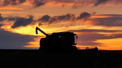 Rob Barrie A combine harvester appears in silhouette on an open field, framed by a dramatic sunset. Orange and pink clouds fill the sky, creating a striking contrast with the dark landscape.