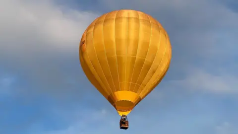 A golden yellow hot air balloon floating in the sky. The sky is blue with a little bit of cloud.