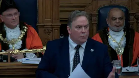 Croydon Council Mayor Jason Perry in a suit and tie with two men behind him. He holds a sheet of paper and stares to the right.