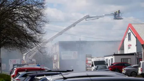 Qays Najm/BBC Firefighters are on a cherry picker-style platform which is on an white mechanical arm leaning across and above an industrial building. The sky is smoky.