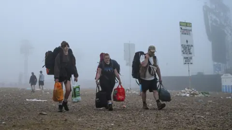 Reuters Three punters leaving Glastonbury Festival. They are all carrying bags and camping gear. There is mist around them and a few piles of rubbish scattered around signs.