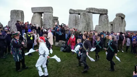 Andrew Matthews/PA Wire Morris dancers in action in front of Stonehenge on a grey day. A crowd watches on, all dressed in winter coats and hats.