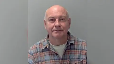 Richard Taylor in a police custody photo. He is wearing a red and blue checked button up shirt over a T-shirt.