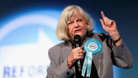 Reuters Ann Widdecombe speaks on the stage during the Reform UK party's rally at the NEC in Birmingham, Britain, June 30, 2024. She is wearing a black and white houndstooth jacket with a large turquoise Reform UK rosette. She is holding a microphone and gesticulating, with a serious expression. Her hair is worn in a blonde bob with a fringe, and she is also wearing some rings on ger fingers and a watch.