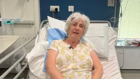 A photo of a woman in a hospital bed, she is wearing a white top with green flowers with some pink on. She is sitting upright and has a little smile. She has white hair. 