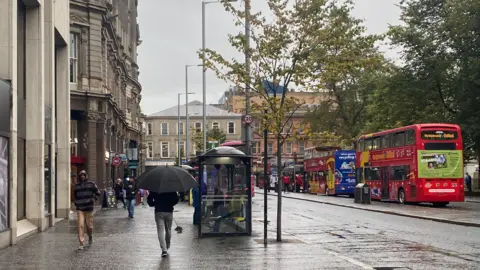 A street in Belfast with red tourist buses are parked and there are people walking along the footpath with umbrellas.