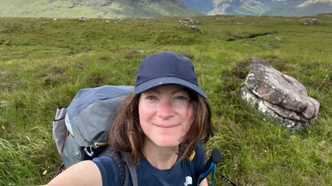 A selfie of Claire Allen standing on a grassy plain with rocks and mountains behind her. She is wearing a blue t-shirt and cap, and is carrying a large backpack