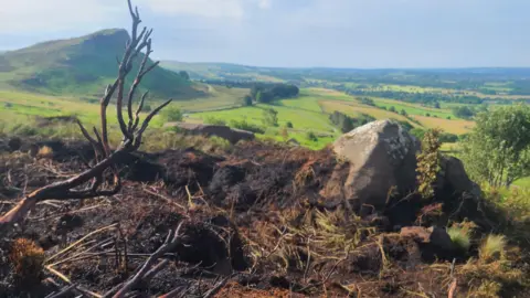 A burnt patch of ground in the middle of the Peak District. A landscape of fields and hills can be seen in the background.