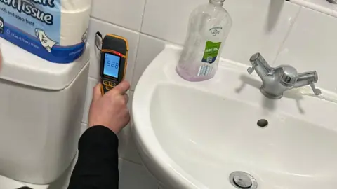 Laura-May A white sink with a metal mixer tap, with white tiles on the wall to the left of the sink and a white toilet system to the left of that. An arm with a black sleeve is visible between the sink and the cistern, and the had is holding a small black and yellow test meter.