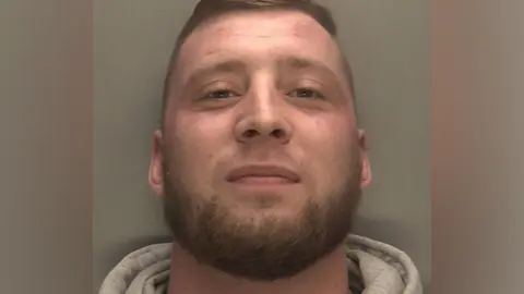 Police mugshot of Adam Shakespeare, who is smiling, wearing a grey jumper with a grey wall in the background