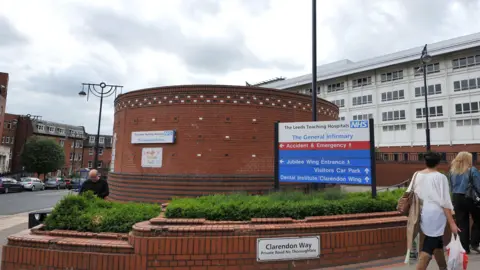 PA Media Main entrance to Leeds General Infirmary - with a circular brick building with a large sign reading Leeds Teaching Hospitals NHS Trust and directions to parts of the site and a large white building behind