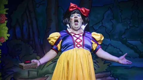 Sheffield Theatres Damian Williams on stage in a yellow and blue dress and black wig in garish makeup