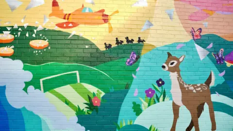 Wychavon District Council Part of a mural on a brick wall. It has rolling hills that are green, with splashing blue waves below it. There are purple butterflies flying and a brown deer in the foreground. The sky is pink and yellow and there is an orange plane flying across it.