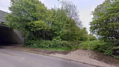The entrance to the proposed housing site in Elsenham. There is a gate across a track into a field, surrounded by trees. A bridge that carries the M11 is on the left.