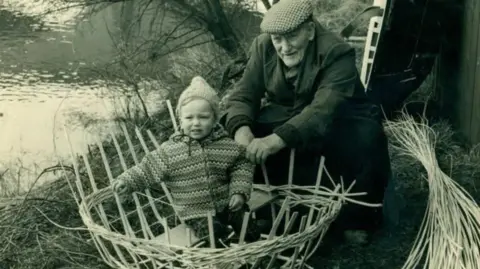 Black and white photo, an elderly man in a flat cap making a round boat out of straw with a baby sat inside along the banks of the river.