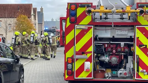 At least ten firefighters can be seen standing to the left of a fire engine. The majority of them are wearing helmets and breathing apparatus. One is pointing off camera at the house that has been damaged.