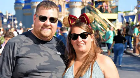 Kevin Clemitson Kevin and Victoria Clemitson are in Disney World. Kevin is on the left wearing a dark grey camouflage style t-shirt and wearing sunglasses. Victoria is on the right wearing Minnie Mouse ears, sunglasses and a blue halter neck top. Rides and other visitors are behind them, out of focus.