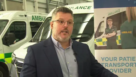 Jon Mawer gives a TV interview in a garage, in front of two ambulances and a company poster. He has greying hair, a close-shaved white beard and glasses and wears an open-necked shirt and dark jacket.