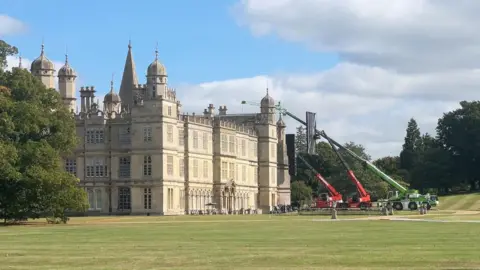 A stately home with cranes holding filming equipment in place for filming. 