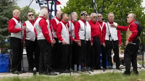 A Gorman Photography A male-voice choir wearing white waistcoats and red shirts sing whilst being conducted by a man wearing a gold an black waistcoat.