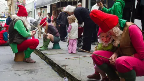 David Chedgey People dressed as gnomes are sat on a street with fishing rods as people watrch on