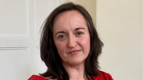A woman with long, dark hair wearing a red top and sitting in a room with a white panelled wall behind her