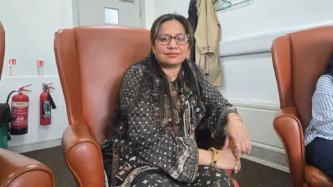Saima is smiling at the camera. She is wearing a dark cotton shirt and has gold cuffs on her wrist with large green stones.