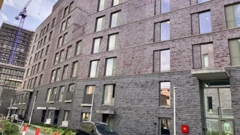 An exterior view of a block of flats. The building is made of grey brick with lots of windows. There are flower beds and parked cars outside the building. 