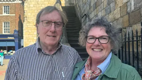 BBC/ Emily Johnson An older man and woman, who both wear glasses, smile in front of a stone staircase leading up to York's city walls.