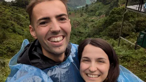 Josh, who has short brown hair and wears a raincoat, smiles in a picture with his fiancee