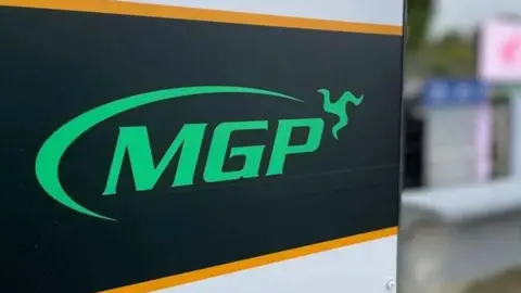 BBC A sign reads MGP in green on a black background; the three legs of man form part of the logo on the top right of the sign.