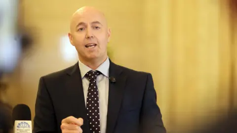 Burrows is wearing a suit and a spotty tie. A staircase is behind him. 