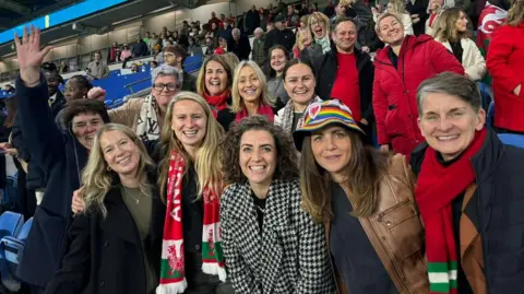 A group of women at a Wales football match wearing hats and scarfs emblazoned with the Welsh badge. There are more fans behind them in the stands.