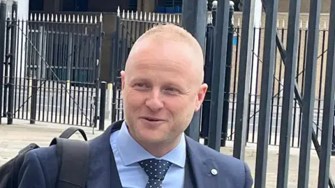 Jamie Bryson leaving court. He has short light hair, wearing a blue suit and carrying a backpack.