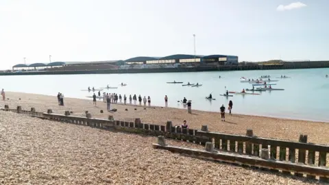 A number of people at Kingston Beach in Shoreham-by-Sea watching a paddleboard vigil. There are people sat on paddleboards out on the water.
