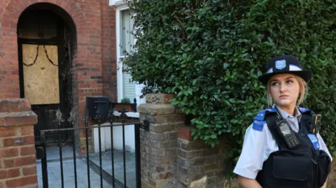 A police officer stands outside a house. A large part of the door is boarded up