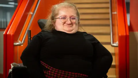 A pistillate   successful  a wheelchair, smiling astatine  the camera. She is wearing a achromatic  apical  with a achromatic  and reddish  skirt successful  a houndstooth pattern. There are stairs down  her with the banisters painted orange. 