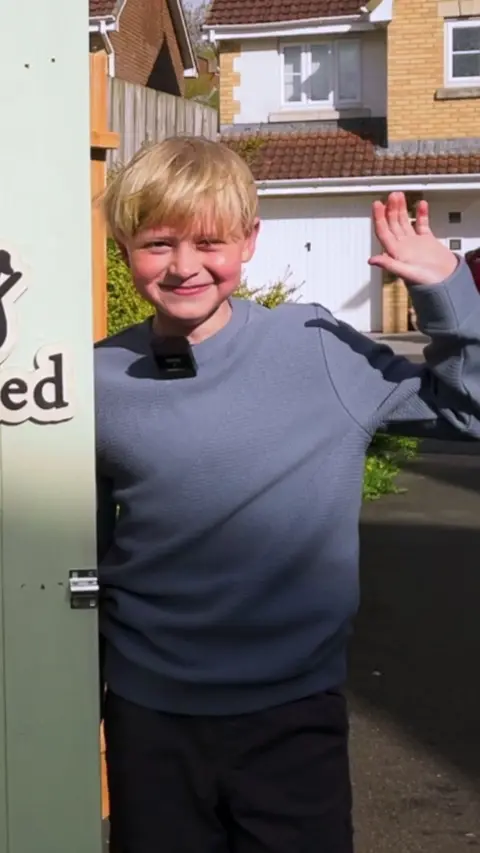 Jacob is wearing a blue jumper and black pants. He is looking at the camera and waving with his left arm. He has blonde hair.
