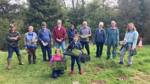 A group of people pose during a planting day at Edensyde in September 2024. There are wearing outdoor gear like wellies. Some are holding up plants. There are nine adults standing in a line in front of trees, some holding garden tools and plants. In front of them is a girl kneeling down and a boy standing up. They both hold black planting trays containing small plants.