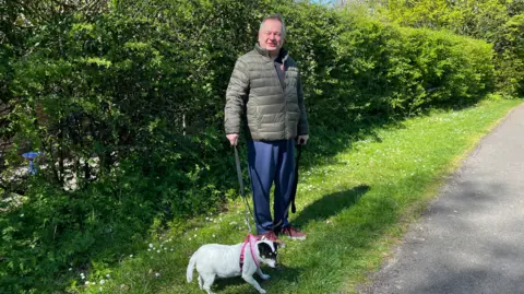 A man is standing on a green grass verge next to a pavement behind a hedge. He is wearing a dark green coat, blue trousers and red shoes. He is holding a lead in his hand, which is connected to a pink harness, with a small Jack Russell dog by his side. She has white fur all over her body, apart from a black and brown face