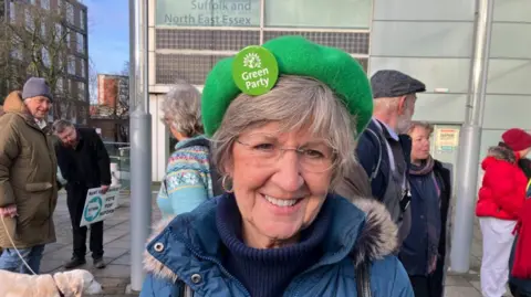 Vikki Irwin/BBC A woman is smiling at the camera and standing outside council offices with a demonstration going on behind her. She is wearing a green hat with a green badge on it which says Green Party. She is also wearing a blue roll neck and with a coat over it. 